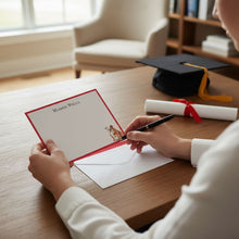 Load image into Gallery viewer, A person is sitting at a desk, writing a personalized graduation note in a red envelope with a pen.